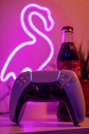 A gaming controller is placed on a surface with a dimly lit neon flamingo sign in the background. Next to the controller is a bottle of Coca-Cola, adding a retro vibe to the composition. The scene is surrounded by a soft pink and purple glow, creating a cozy and nostalgic atmosphere.