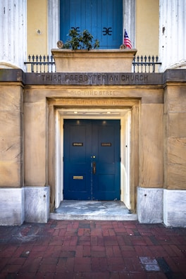 The entrance to a historical church with wooden doors painted in blue. Above the doors, there is a sign indicating 'Third Presbyterian Church' and an address on Old Pine Street. The building is constructed with stone and features white columns on either side. An American flag is visible above the entrance.