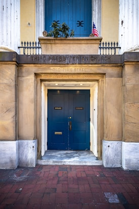 The entrance to a historical church with wooden doors painted in blue. Above the doors, there is a sign indicating 'Third Presbyterian Church' and an address on Old Pine Street. The building is constructed with stone and features white columns on either side. An American flag is visible above the entrance.