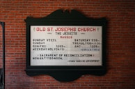 A church sign is mounted on a brick wall, displaying the service times for Old St. Joseph's Church. The sign includes information about the Jesuits and their Mass schedules, including times for Sunday Vigil, Sunday, Monday to Friday, and weekday holydays. It also mentions the Sacrament of Reconciliation with available times.