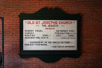 A church sign is mounted on a brick wall, displaying the service times for Old St. Joseph's Church. The sign includes information about the Jesuits and their Mass schedules, including times for Sunday Vigil, Sunday, Monday to Friday, and weekday holydays. It also mentions the Sacrament of Reconciliation with available times.