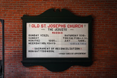 A church sign is mounted on a brick wall, displaying the service times for Old St. Joseph's Church. The sign includes information about the Jesuits and their Mass schedules, including times for Sunday Vigil, Sunday, Monday to Friday, and weekday holydays. It also mentions the Sacrament of Reconciliation with available times.