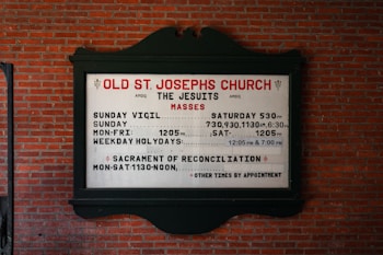 A church sign is mounted on a brick wall, displaying the service times for Old St. Joseph's Church. The sign includes information about the Jesuits and their Mass schedules, including times for Sunday Vigil, Sunday, Monday to Friday, and weekday holydays. It also mentions the Sacrament of Reconciliation with available times.