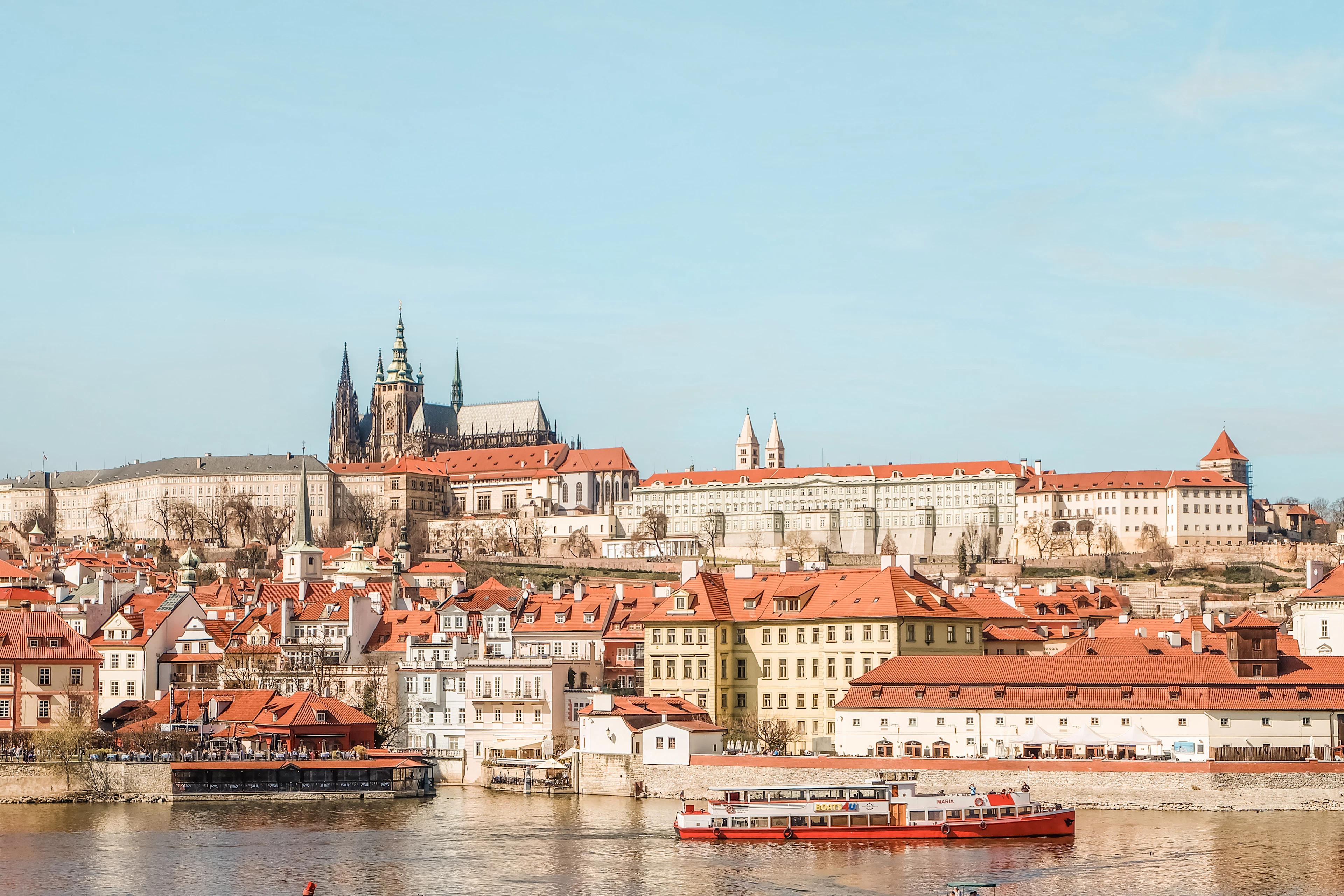 Prague Castle and St. Vitus Cathedral rise above the Vltava River, boats passing beneath red rooftops.
