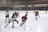 A group of young hockey players, wearing gear and uniforms, are actively engaged in a game on an indoor ice rink. The players are from opposing teams, one in red and the other in blue and white, and they are focused on the puck. The background shows the hockey rink's boards and a referee overseeing the match.