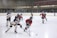 A group of young hockey players, wearing gear and uniforms, are actively engaged in a game on an indoor ice rink. The players are from opposing teams, one in red and the other in blue and white, and they are focused on the puck. The background shows the hockey rink's boards and a referee overseeing the match.