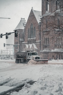 A snowplow truck is clearing a snow-covered street in front of a large, historical stone building that resembles a church. Snow is falling heavily, creating a wintry scene. The building features arched windows and detailed stonework. Traffic lights are visible, and a tree on the right adds to the urban winter landscape.