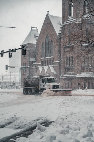 A snowplow truck is clearing a snow-covered street in front of a large, historical stone building that resembles a church. Snow is falling heavily, creating a wintry scene. The building features arched windows and detailed stonework. Traffic lights are visible, and a tree on the right adds to the urban winter landscape.