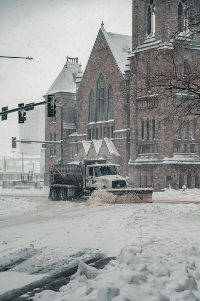 A snowplow truck is clearing a snow-covered street in front of a large, historical stone building that resembles a church. Snow is falling heavily, creating a wintry scene. The building features arched windows and detailed stonework. Traffic lights are visible, and a tree on the right adds to the urban winter landscape.