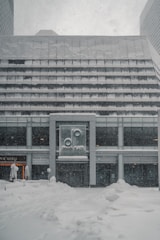 A modern building exterior covered in a heavy blanket of snow. The signage on the front displays the name 'Denver Place.' Snowflakes are visibly falling, suggesting an ongoing snowstorm. The ground is entirely covered in thick snow, adding to the wintry atmosphere.