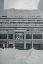 A modern building exterior covered in a heavy blanket of snow. The signage on the front displays the name 'Denver Place.' Snowflakes are visibly falling, suggesting an ongoing snowstorm. The ground is entirely covered in thick snow, adding to the wintry atmosphere.