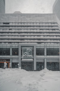 A modern building exterior covered in a heavy blanket of snow. The signage on the front displays the name 'Denver Place.' Snowflakes are visibly falling, suggesting an ongoing snowstorm. The ground is entirely covered in thick snow, adding to the wintry atmosphere.