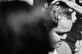 A black and white photograph of a person having their hair cut. A hand is placed on top of the person’s head, guiding the process. The lighting creates strong contrasts, highlighting the facial features of the individual.
