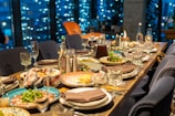 A dining table is elegantly set with diverse plates of food, including salads and entrees. Crystal glasses and cutlery are arranged alongside pitchers of beverages. The background features a window with a view of city lights, creating a cozy and sophisticated atmosphere.