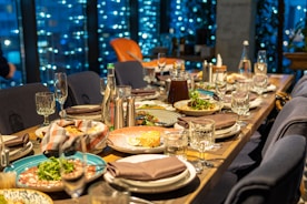 A dining table is elegantly set with diverse plates of food, including salads and entrees. Crystal glasses and cutlery are arranged alongside pitchers of beverages. The background features a window with a view of city lights, creating a cozy and sophisticated atmosphere.