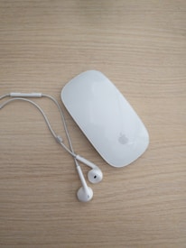 A white wireless computer mouse and a pair of white earphones rest on a light wooden surface. The mouse is sleek and glossy, with a minimalist design. The earphones are untangled, positioned next to the mouse, with their wires neatly laid out.