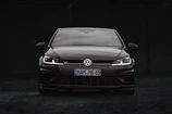 A sleek black Volkswagen car is positioned at the center of the image. The vehicle is viewed from the front, showcasing its grille, headlights, and the iconic Volkswagen logo. The headlights are on, creating a striking contrast against the dark background.