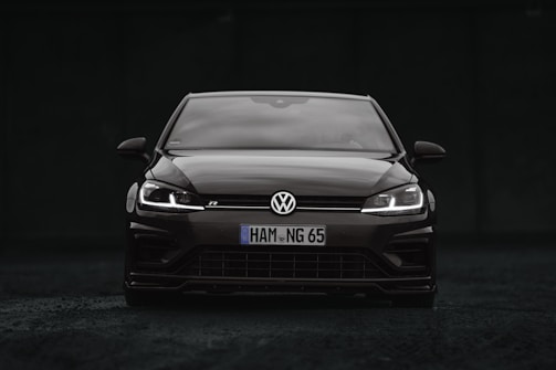 A sleek black Volkswagen car is positioned at the center of the image. The vehicle is viewed from the front, showcasing its grille, headlights, and the iconic Volkswagen logo. The headlights are on, creating a striking contrast against the dark background.