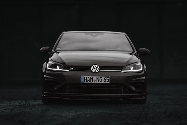A sleek black Volkswagen car is positioned at the center of the image. The vehicle is viewed from the front, showcasing its grille, headlights, and the iconic Volkswagen logo. The headlights are on, creating a striking contrast against the dark background.