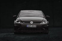A sleek black Volkswagen car is positioned at the center of the image. The vehicle is viewed from the front, showcasing its grille, headlights, and the iconic Volkswagen logo. The headlights are on, creating a striking contrast against the dark background.