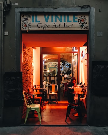 The entrance of a cozy caf&eacute; bar named 'Il Vinile' is warmly illuminated with orange lights. Inside, there's a mix of retro and modern decor with vinyl records displayed on the walls. A woman is seated at a table, reading or writing under the inviting light. The atmosphere suggests a relaxed and intimate setting.