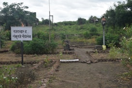 A garden path is bordered by stones and surrounded by lush greenery. There is a sign with text in a script that appears to be Marathi. The path leads towards a small stack of bricks. In the background, there are trees and a slightly overcast sky.
