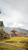 A traditional Balinese temple structure surrounded by well-maintained gardens with hedges and grass. The temple features typical architecture with a tall, triangular roof. In the background, a lush green mountain is partially covered by clouds, adding a dramatic effect to the scene. Several people are visible walking around, suggesting it is a popular tourist destination.