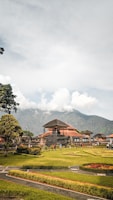 A traditional Balinese temple structure surrounded by well-maintained gardens with hedges and grass. The temple features typical architecture with a tall, triangular roof. In the background, a lush green mountain is partially covered by clouds, adding a dramatic effect to the scene. Several people are visible walking around, suggesting it is a popular tourist destination.