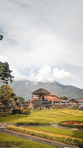 A traditional Balinese temple structure surrounded by well-maintained gardens with hedges and grass. The temple features typical architecture with a tall, triangular roof. In the background, a lush green mountain is partially covered by clouds, adding a dramatic effect to the scene. Several people are visible walking around, suggesting it is a popular tourist destination.