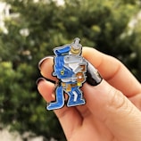 Close-up of a hand holding a cartoon-themed enamel pin of a blue-clad character in a police outfit, complete with a hat and belt, holding a coffee and donut. The background is blurred green foliage.