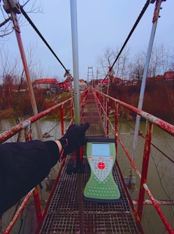 A long suspension bridge stretches over a body of water, with rusted red railings and a grid walkway. A person holding surveying equipment stands on the bridge. Bare trees line the water's edge, and buildings with red roofs are visible in the background under an overcast sky.