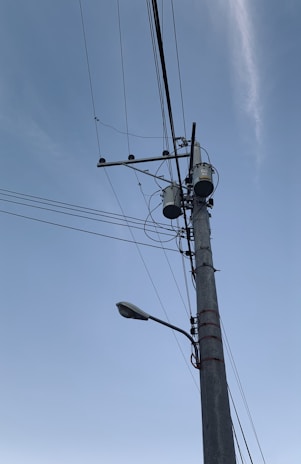 A tall utility pole is equipped with multiple wires and two large cylindrical transformers. A streetlamp extends outward from the pole, which is set against a clear blue sky.