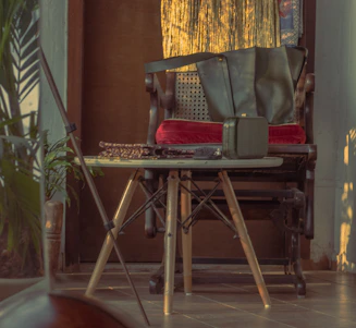 A sunlit corner featuring a vintage pine writing desk beside a padded fabric accent chair, with woven rope plant holders hanging nearby.