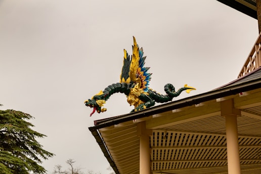 A colorful dragon sculpture is perched on the corner of a roof. The dragon has vibrant blue, yellow, and green scales with an elaborate design. Its wings are spread wide, displaying bright feathers, while its mouth is open, showing sharp teeth and a red tongue. The roof beneath is made of wood with a beige trim, and the sky in the background is overcast.