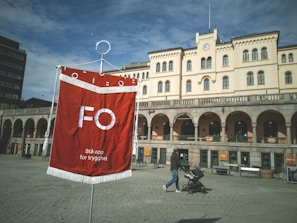 A red banner with white text and fringes is prominently displayed in front of a historic building with arched windows and a clock tower. A person pushes a stroller in the foreground, while the backdrop is a clear, blue sky.