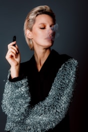 A person with short hair is holding a vape pen and exhaling smoke, wearing a black coat with textured, reflective details on the sleeves. The background is a solid, muted color, enhancing the focus on the person's contemplative expression and the soft wisp of smoke.