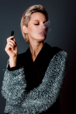 A person with short hair is holding a vape pen and exhaling smoke, wearing a black coat with textured, reflective details on the sleeves. The background is a solid, muted color, enhancing the focus on the person's contemplative expression and the soft wisp of smoke.