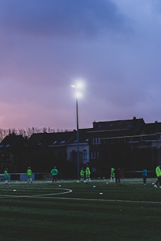 A group of people wearing bright green sports attire can be seen on a sports field during dusk. The field is well-lit by a high floodlight, and a row of houses is visible in the background under a purple sky.