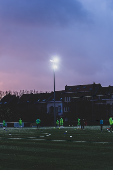 A group of people wearing bright green sports attire can be seen on a sports field during dusk. The field is well-lit by a high floodlight, and a row of houses is visible in the background under a purple sky.