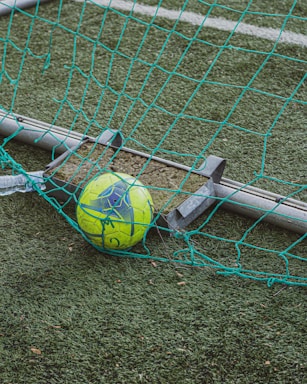 A bright yellow and blue soccer ball is trapped in a green net on a grass football field. The net appears slightly damaged and is positioned on a metal frame. The artificial turf has a realistic texture with visible white boundary lines in the background.