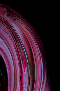 A close-up abstract image of swirling lines and vibrant colors resembling fluid art or marbled patterns. The dominant hues are pink, red, and blue, set against a dark, nearly black background. The image captures a sense of motion and fluidity through its curved lines and blending colors.
