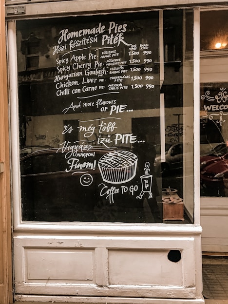 A shop window displays a menu for homemade pies, written in both English and another language, possibly Hungarian. The menu lists various pie flavors such as Spicy Apple Pie and Chicken Liver Pie, along with their prices. There are also decorative drawings of a pie and a cup of coffee with the text 'Coffee To Go.' The overall design is done in white chalk on a dark window background.