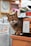 A tabby cat sits on the edge of a counter next to a jar labeled with a price tag written in both English and a non-English script. The background features various items, suggesting a cozy, possibly cluttered shop environment. A blurred clock is visible, adding a sense of time to the setting.