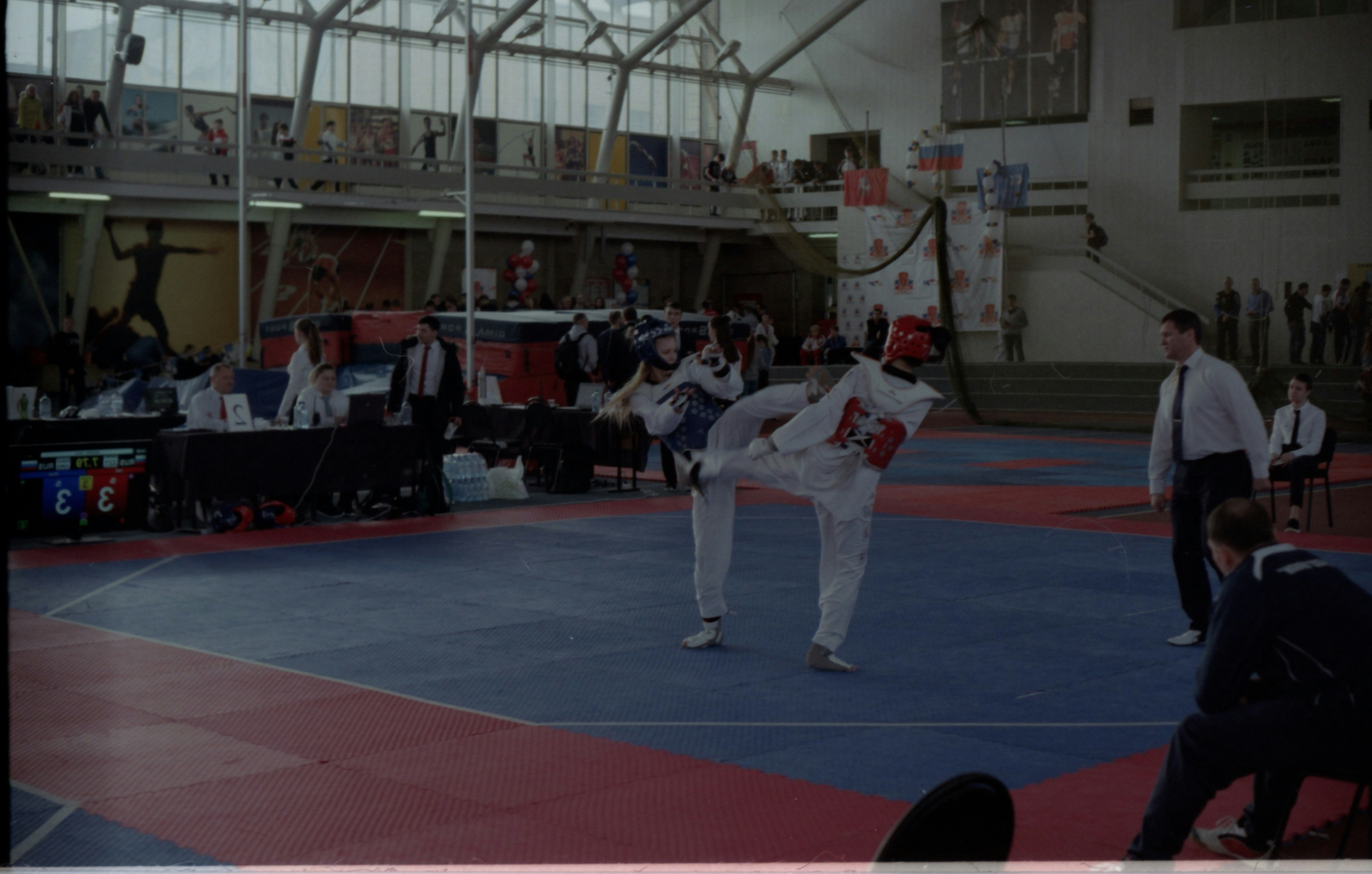 Two advanced taekwondo sparring athletes in red and blue gear mid-fight in dark atmospheric competition hall