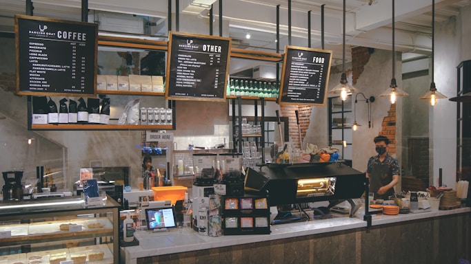A modern coffee shop interior with an industrial design featuring exposed brick, metal shelving, and pendant lighting. Menu boards hang from the ceiling above a counter displaying coffee and food options. A barista stands behind the counter with a large espresso machine, and various containers and tools for coffee preparation are visible. Shelves hold coffee bags and packaged goods, while a display case contains pastries.