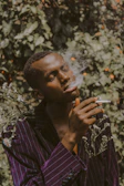 A person with closed eyes gently exhales smoke while holding a smoking item in hand. The background features leafy plants, and the individual is wearing a dark, pinstriped suit adorned with small white flowers.