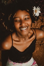 A smiling woman with eyes closed, wearing a black lace top. Her hair is styled naturally with a white daisy flower tucked into it. The background is earthy and natural.