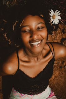A smiling woman with eyes closed, wearing a black lace top. Her hair is styled naturally with a white daisy flower tucked into it. The background is earthy and natural.