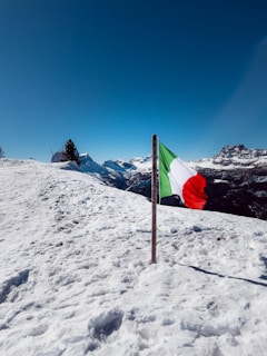 A snowy landscape with a clear blue sky and mountain peaks in the background. An Italian flag is prominently displayed, erected on a pole in the snow.