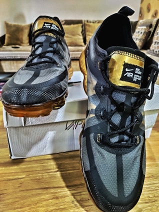 A pair of athletic shoes with a sleek, modern design featuring a black and gray color scheme with gold accents. The shoes are placed on a wooden surface, with a white shoe box in the background. The laces are black, and the shoes have a transparent, amber-colored sole.